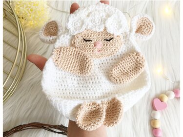 Gehäkeltes weiß-beiges Schaf-Schmusetuch mit schlafendem Gesicht, in einer Hand gehalten vor hellem Hintergrund.