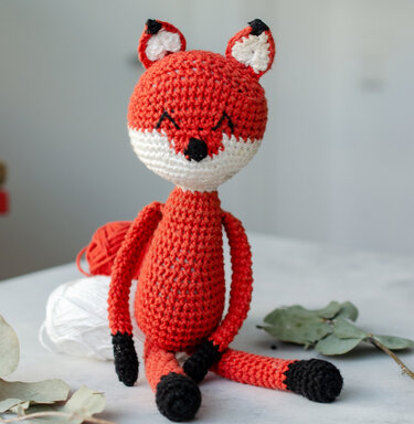 Sitzender Amigurumi-Fuchs aus orangefarbener Wolle mit Garn im Hintergrund