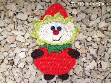Draufsicht einer gehäkelten Erdbeer-Applikation mit Gesicht und grünen Blättern auf hellem Kies