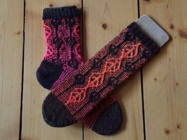 Zwei gestrickte Socken mit Blattmuster in Pink und Orange auf Holzdielen