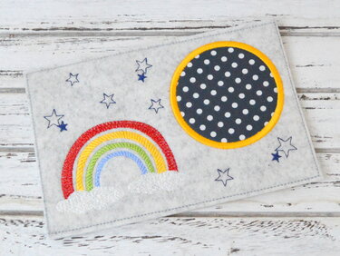Hellgrauer Filz-Tassenteppich mit gesticktem Regenbogen, Wolken, Sternen und dunkelblauem Punkte-Kreis mit gelbem Rand.