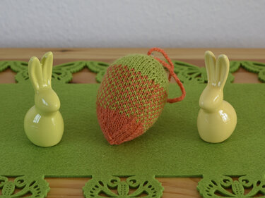 Gestricktes Ei zwischen zwei hellgelben Hasenfiguren auf grünem Filz
