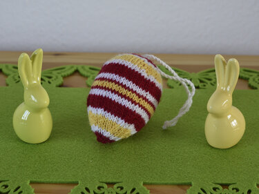 Gestricktes gestreiftes Osterei in Rot, Weiß und Gelb mit weißer Schnur, flankiert von zwei gelben Keramikhasen auf grüner Filzmatte