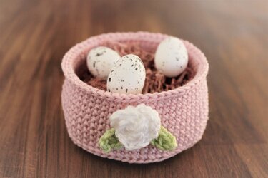 Rosa gehäkelter Korb mit drei gesprenkelten Eiern und weißer Blüte.