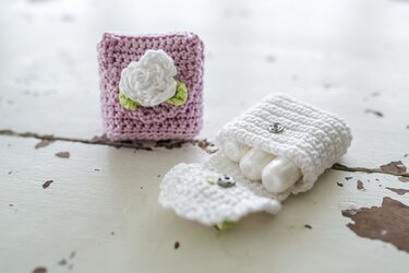 Zwei gehäkelte Täschchen: ein rosa mit weißer Häkelrose, das andere weiß und geöffnet mit drei Tampons, auf einem alten Holztisch.