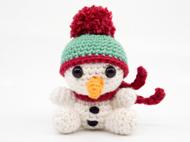 White crocheted amigurumi snowman wearing a green hat with red trim, a red scarf, orange nose and black eyes.