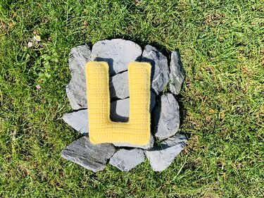Gelber, gehäkelter Buchstabe U auf grauen Steinen im grünen Gras.