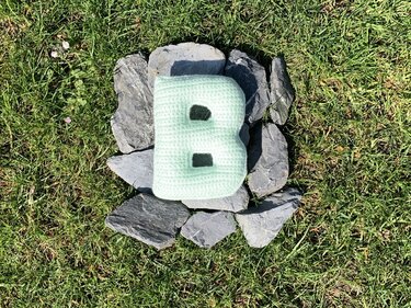 Gehäkeltes hellmintfarbenes Kissen in Form des Buchstabens B auf Schiefersteinen im Rasen