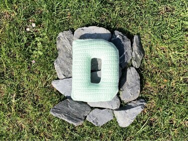 Nahaufnahme eines gefüllten, gehäkelten Kissens in Form des Buchstabens D auf Schiefersteinen