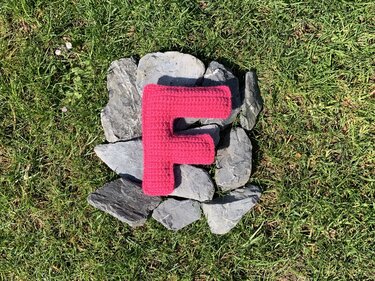 Gehäkelter Buchstabe F in Pink auf Grausteinen im grünen Rasen