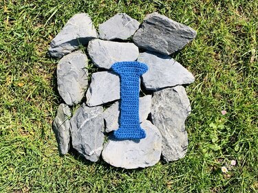 Gehäkelter blauer Buchstabe I auf grauen Steinen im Gras