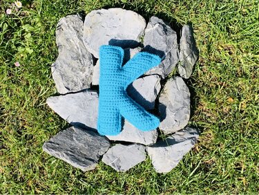 türkis gehäkelter Buchstabe K auf flachen Steinen im Gras