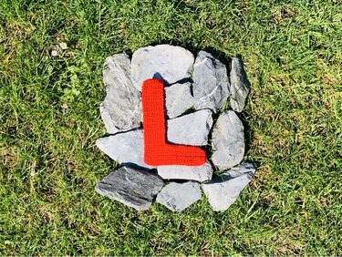 Roter gehäkelter Buchstabe L auf grauen Steinen im Gras