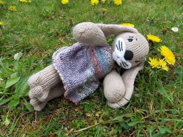 Beiges gestricktes Kaninchen-Amigurumi mit bunter Weste im Gras, umgeben von Löwenzahn