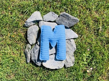 Gehäkelter blauer Buchstabe M auf grauen Steinen in grünem Gras