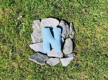 Hellblauer gehäkelter Buchstabe N auf grauen Schieferplatten im Gras