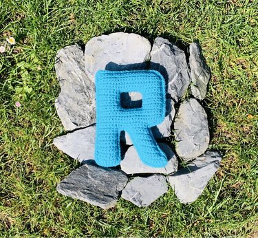 Gehäkelter Buchstabe R (türkis) auf Gras, umgeben von Steinen