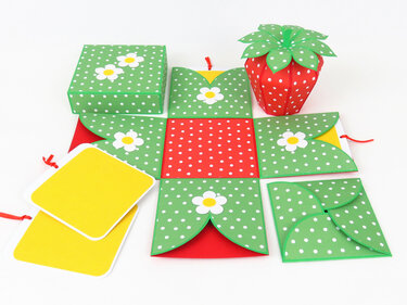 Geöffnete grüne gepunktete Explosionsbox mit weißen Blumenmotiven, rotem Zentrum, gelben Einlegerkarten und einer erdbeerförmigen roten Geschenkbox