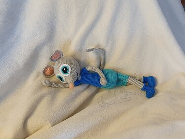 Gehäkelte graue Amigurumi‑Maus mit großen türkisfarbenen Augen, blauem Oberteil und türkiser Hose liegend auf heller Decke