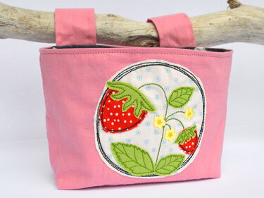 Rosa Stofftasche mit runder Erdbeer-Applikation mit weißen Punkten, grünen Blättern und gelben Blüten, an Treibholz hängend.