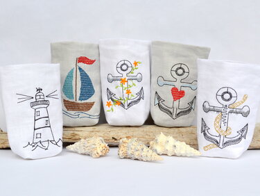 Fünf bestickte Stoffhüllen mit maritimen Motiven (Leuchtturm, Segelboot, Anker) für LED‑Tischlichter auf Treibholz mit Muscheln