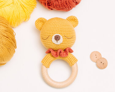 Yellow crocheted bear baby rattle with orange ruffle and natural wooden ring, shown with yarn and wooden buttons.