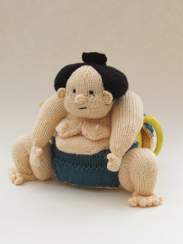 Knitted sumo wrestler tea cozy in beige with black hair and teal belt on a teapot