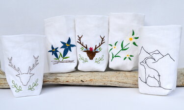 Fünf weiße Stoffhüllen mit gestickten alpinen Motiven (Hirschkopf, Enzian, Hirsch mit Blumen, Edelweiß, Steinbock) auf einem Holzstamm.