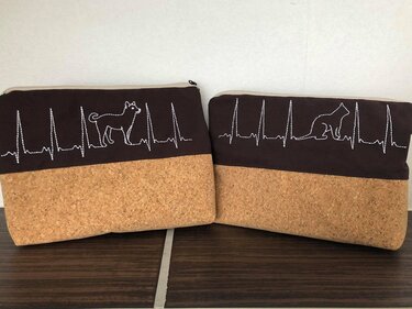 Nahaufnahme zweier Täschchen mit Korkboden und EKG-Stickmotiv; links Hundesilhouette, rechts Katzensilhouette
