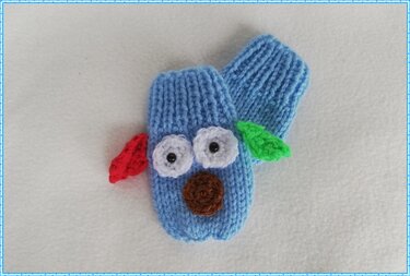 Zwei hellblaue gestrickte Babyfäustlinge mit gehäkelten weißen Augen, schwarzer Mitte, brauner Nase und roten sowie grünen seitlichen Applikationen auf weißem Hintergrund