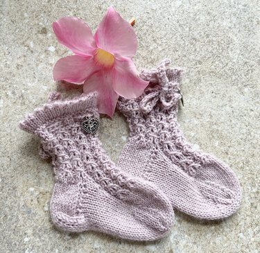 Zwei handgestrickte Babysocken in Lavendelfarben mit Rüschenbündchen und Herzanhänger neben einer rosa Blüte auf hellem Untergrund.