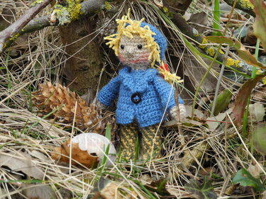 Gehäkelte Puppe mit blauer Jacke und gelbem Haar im Gras neben Zapfen