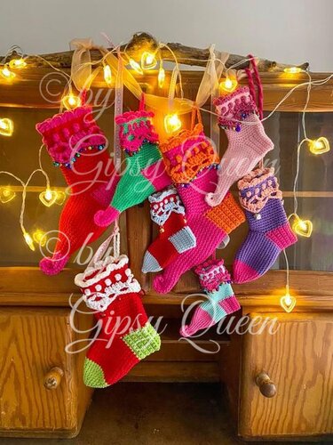 Bunte gehäkelte Mini-Weihnachtsstiefel mit Perlen und Bändern an einem Ast vor einem Holzschrank und herzförmigen Lichterketten