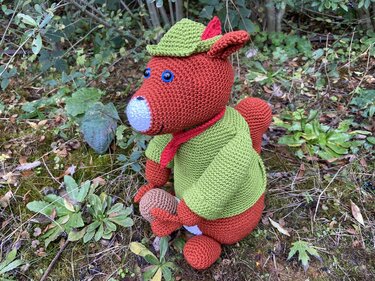 Rotbraunes gehäkeltes Eichhörnchen-Amigurumi mit grüner Jacke, grünem Hut mit roter Feder und rotem Halstuch hält eine Nuss auf moosigem Waldboden.