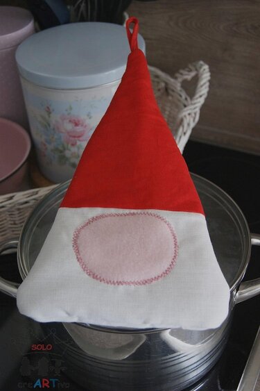 Ein rot-weißer Topflappen im Zwergenmützen-Design mit einer rosa ovalen Applikation, der auf einem Kochtopf liegt.