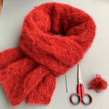 Kuscheliger roter Strickschal mit Stopfnadel, kleiner roter Schere und Garnrest auf neutralem Hintergrund.