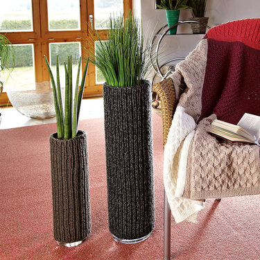 Zwei hohe Glasvasen mit dunkelgrauen gestrickten Überzügen auf rotem Teppich neben einem Sessel mit Strickdecke