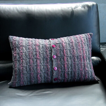 Gestricktes rechteckiges Kissen grau-rosa mit drei pinken Knöpfen auf schwarzer Couch