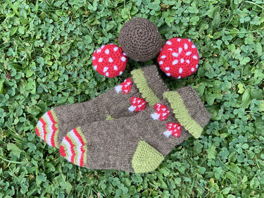 Gestrickte braune Babysocken mit grünen Bündchen und roten Pilz‑Applikationen, dazu zwei gehäkelte rot‑weiße Pilze auf Kleewiese