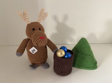 Gehäkeltes Rentier mit Schild „21“ neben Häkelkorb mit bunten Kugeln und grünem Häkelbaum.