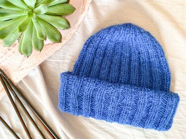Nahaufnahme einer blau gestrickten Rippmütze mit umgeschlagenem Bund, Stricknadeln liegen daneben
