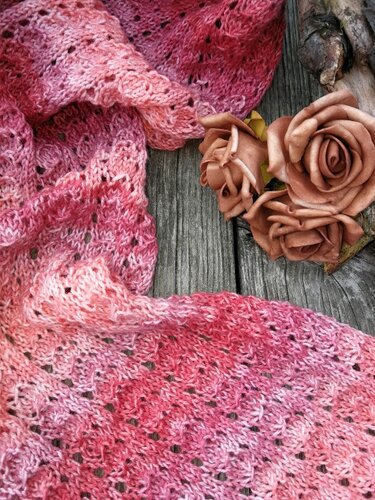 Gestrickter rosa Ajour-Schal mit Farbverlauf auf Holzbord, daneben dekorative Rosen