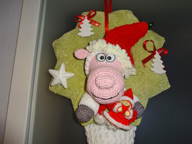 Gehäkeltes Schaf mit Weihnachtsmütze und Holzengel auf grünem Sternkissen, umgeben von Tannen- und Sternanhängern.