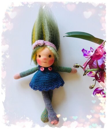 Gehäkelte Amigurumi-Puppe mit grünem Haar, blauem Kleid und rosa Stirnband neben violetten Orchideen
