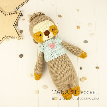 Gehäkeltes Amigurumi-Faultier mit blauem Pullover und rosafarbenem Herz, liegend auf hellem Holz