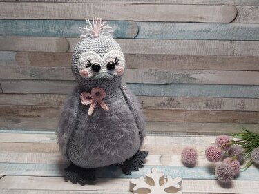 Gehäkeltes graues Amigurumi-Vögelchen mit flauschigen Flügeln, großen schwarzen Augen und rosafarbener Schleife vor Holzpaneelen