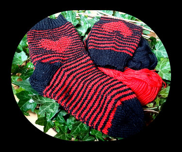 Rot-schwarz gestreifte Socken mit Herz am Bündchen neben Garn und Stricknadeln auf Efeublättern.