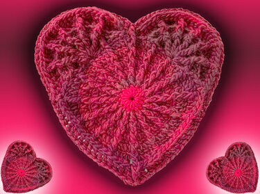 Gehäkeltes Herz in Rosa- und Rottönen mit Spiralmuster, zwei kleinere Herzen unten, verlaufender pinker Hintergrund