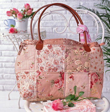 Große gesteppte Einkaufstasche mit floralen Patchwork-Quadraten und braunen Henkeln auf einem weißen Stuhl