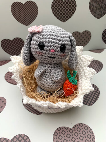 Grauer Amigurumi-Häkelhase mit rosa Blüte im weißen Häkelkörbchen auf Stroh, daneben eine kleine gehäkelte Karotte.
